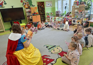 Zdjęcie przedstawia dzieci siedzące w kole na dywanie oraz mamę Tosi czytającą książeczkę.