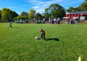 Zdjęcie przedstawia dzieci podczas zabaw sportowych - prowadzenie nogą piłki do pachołka.
