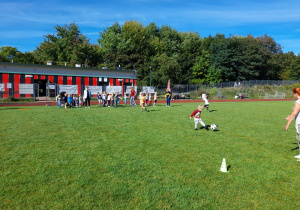 Zdjęcie przedstawia dzieci podczas zabaw sportowych - prowadzenie nogą piłki do pachołka.