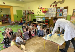 Zdjęcie przedstawia dzieci i prowadzącego zajęcia, prezygotowujacego się do wykonywania eksperymentów.
