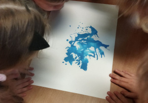 Zdjęcie przedstawia dzieci wykonujące zadanie - rozdmuchiwanie kropli niebieskiej farby.