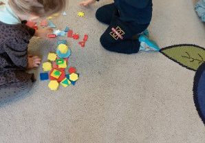 Zdjęcie przedstawia dzieci z grupy I podczas zabaw z figurami geometrycznymi na dywanie.