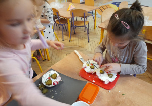 Na tym zdjęciu widać dziewczynki, które układają pokrojone w plasterki warzywa na bułce, tworząc oczy, uszy, nos i wąsiki.