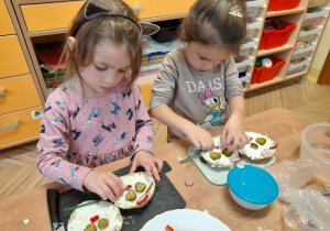Na tym zdjęciu widać dziewczynki, które układają pokrojone w plasterki warzywa na bułce, tworząc oczy, uszy, nos i wąsiki.