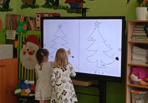 Zdjęcie przedstawia dziewczynki rysujące ozdoby na choince przy tablicy interaktywnej.