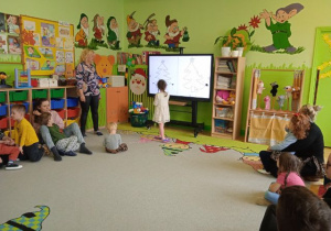 Zdjęcie przedstawia dzieci z rodzicami oraz dziewczynkę rysującą ozdoby na choince przy tablicy interaktywnej.
