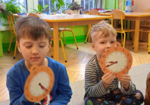 Zdjęcie przedstawia Tymona z grupy II oraz Adasia z grupy I podczas zabawy utrwalającej cyfry na tarczy zegara.