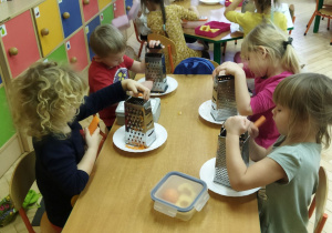 Zdjęcie przedstawia dzieci, które scierają na tarkach marchewki.