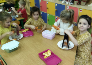 Zdjęcie przedstawia dzieci scierające marchewki na tarkach.