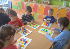 Zdjęcie przedstawia dzieci z grupy I i II podczas gry planszowej z wykorzystaniem kolorowych klocków plastykowych.