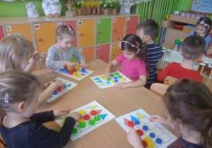 Zdjęcie przedstawia dzieci z grupy I i II podczas gry planszowej z wykorzystaniem kolorowych klocków plastykowych.