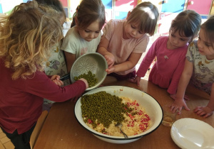 Zdjęcie przedstawia dzieci przesypujące groszek do miski z innymi składnikami.