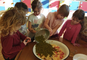 Przedstawia dzieci przesypujące groszek do miski z innymi składnikami.
