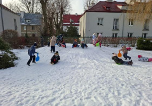 Zdjęcie przedstawia dzieci bawiące się na zaśnieżonej górce oraz zjeżdżajace z niej na jabłuszkach.