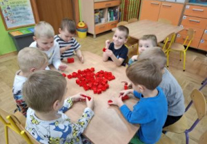 Zdjęcie przedstawia dzieci z grupy II podczas zabawy przy stolikach z wykorzystaniem kolorowych klocków.