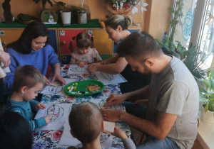 Na tym zdjęciu widać troje dzieci wraz z rodzicami podczas wykonywania pracy plastycznej.