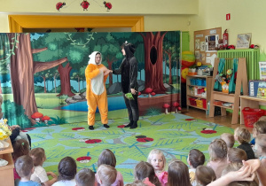 Zdjęcie przedstawia jedną ze scen teatrzyku oraz dzieci oglądające przedstawienie.