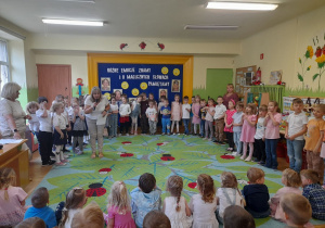 Zdjęcie przedstawia powitanie młodszych dzieci przez nauczycielkę oraz starsze dzieci przygotowane do występu.
