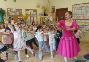 Zdjęcie przedstawia dzieci, które pod okiem baletnicy wykonują figury baletowe.