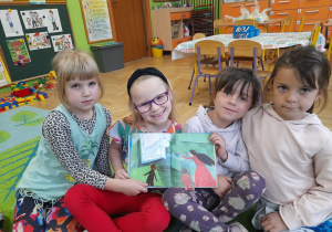 Zdjęcie przedstawia Wandę z grupy III oraz Oliwię, Hanię T., Marysię z grupy IV prezentujące jedną z ilustracji w czytanej książeczce.