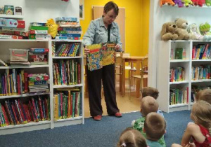 Zdjęcie przedstawia panią bibliotekarkę czytającą dzieciom bajkę i pokazującą ilustracje oraz słuchające dzieci.