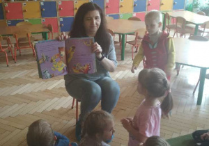 Zdjęcie przedstawia panią bibliotekarkę, która pokazuje dzieciom ilustracje w książce.