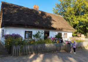 Zdjęcie przedstawia dzieci przed wejściem do starej, wiejskiej chaty.