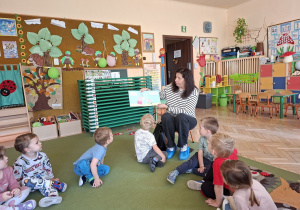 Zdjęcie przedstawia kilkoro dzieci słuchających bajki czytanej przez panią bibliotekarkę.