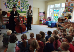 Zdjęcie przedstawia dzieci podczas prezentacji kontrabasu przez kontrabasistkę oraz prowadzącego.