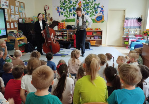 Zdjęcie przedstawia dzieci podczas prezentacji kontrabasu przez kontrabasistkę oraz prowadzącego.
