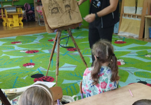 Zdjęcie przedstawia ilustratora przy sztaludze, na której szkicował węglem rysunki.