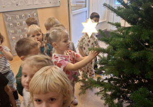 Zdjęcie przedstawia dzieci z grupy I przy choince i Kamilkę wieszajacą bombkę.