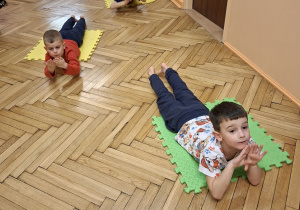 Zdjęcie przedstawia trzech chłopców z grupy III podczas wykonywania ćwiczeń na matach w leżeniu przodem.