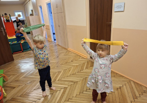 Zdjęcie przedstawia Natalkę i Damiana z grupy I wykonujących ćwiczenia rozciągające mięśnie klatki piersiowej z wykorzystaniem szarf.