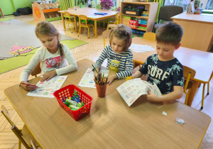 Zdjęcie przedstawia Klarę J., Julka oraz Stasia z grupy IV kolorujących i wycinających elementy wyposażenia lodówki.