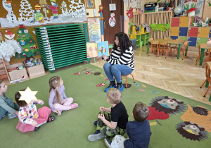 Na tym zdjęciu widać kilkoro dzieci oraz panią bibliotekarkę, która czyta dzieciom książkę.
