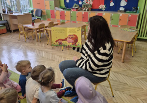 Zdjęcie przedstawia panią bibliotekarkę, która czyta dzieciom książkę przyniesioną z bilblioteki.