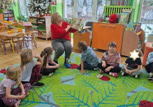 Zdjęcie przedstawia panią bibliotekarkę czytającą książkę oraz słuchające opowiadania dzieci.
