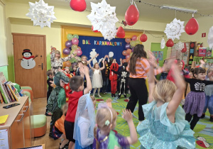 Na tym zdjęciu widać dzieci biorące udział w zabawach karnawałowych przy muzyce prowadzonych przez wodzirejkę.