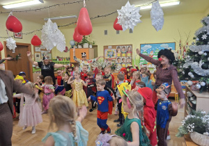 Na tym zdjęciu widać dzieci biorące udział w zabawach karnawałowych przy muzyce prowadzonych przez wodzirejkę.