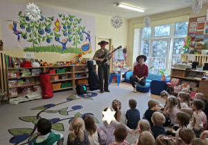 Zdjęcie przedstawia dzieci słuchające gry na gitarze w wykonaniu jednego z muzyków.