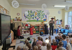 Zdjęcie przedstawia dzieci słuchające gry na gitarze w wykonaniu jednego z muzyków.
