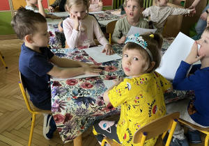 Zdjęcie przedstawia dzieci siedzące przy stolikach wykonujące kartę pracy.