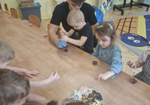 Zdjęcie przedstawia Mikołaja L. napełniającego foremkę płynną czekoladą oraz Apolonię i inne dzieci ozdabiajace swoje czekoladki.