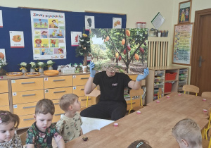 Na tym zdjęciu widać prowadzącego zajęcia, który prezentuje dzieciom planszę przedstawiającą drzewo kakaowca.