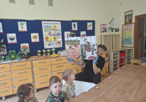 Zdjęcie przedstawia dzieci oraz prowadzącego, który prezentuje planszę przedstawiającą proces powstawania czekolady.