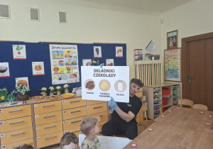 Na tym zdjęciu widać prowadzącego przedstawiającego dzieciom ilustrację przedstawiającą składniki czekolady.