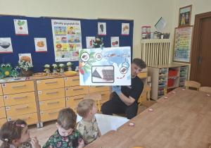 Zdjęcie przedstawia prowadzacego zajęcia, który opowiada dzieciom jak powstaje czekolada i ilustruje ten proces obrazkami.