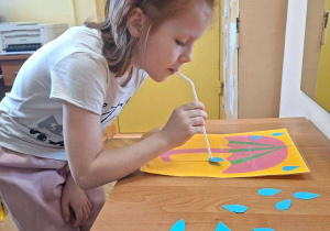 Zdjęcie przedstawia Marcelinę T. z grupy IV wykonującą ćwiczenie oddechowe - przenoszenie papierowych kropelek deszczu na parasol za pomoca słomki do napojów.