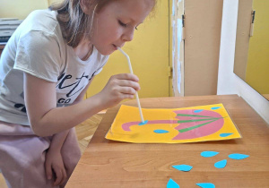 Zdjęcie przedstawia Marcelinę T. z grupy IV wykonującą ćwiczenie oddechowe - przenoszenie papierowych kropelek deszczu na parasol za pomoca słomki do napojów.